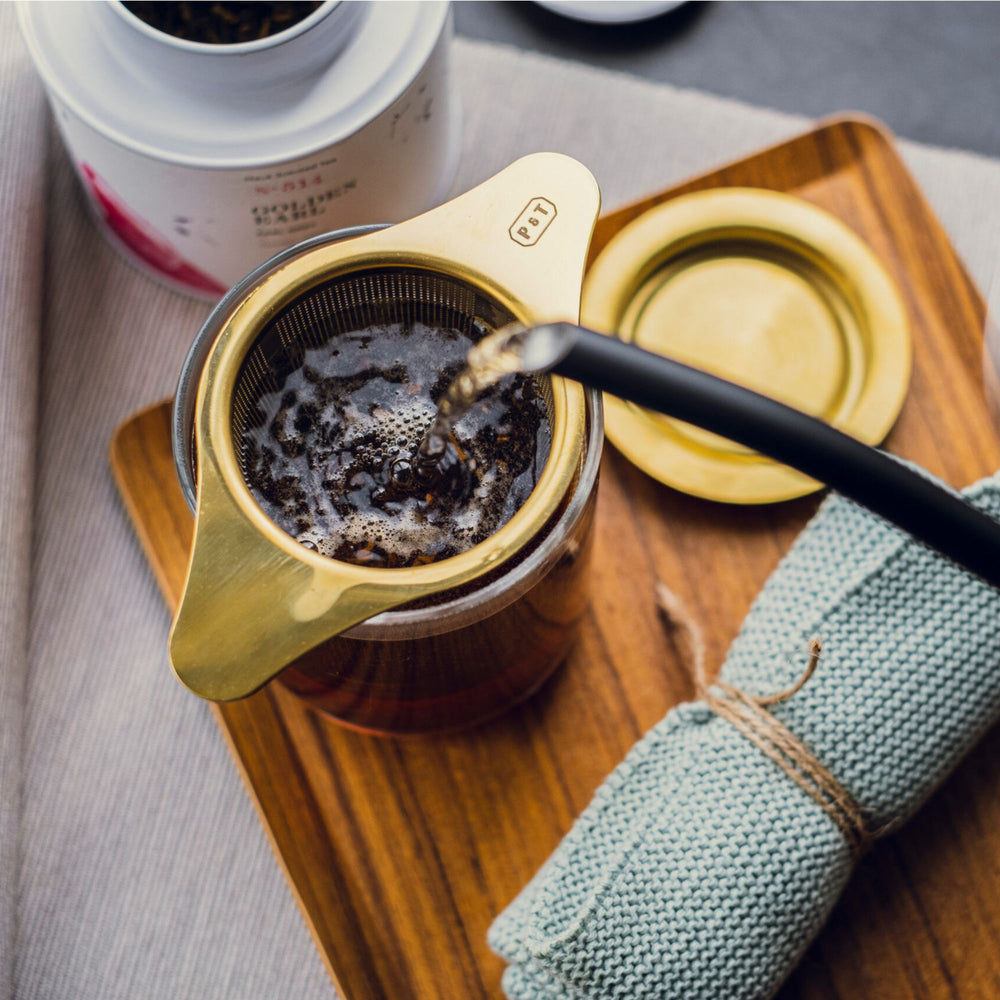 Glas-Kaffeebereiter mit goldenem Filter auf Holztablett, umgeben von Stricktuch, Deckel und Behälter auf Leinentuch.