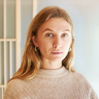 Junge Frau mit langen blonden Haaren und grünen Augen im beige gerippten Pullover mit goldenen Creolen.