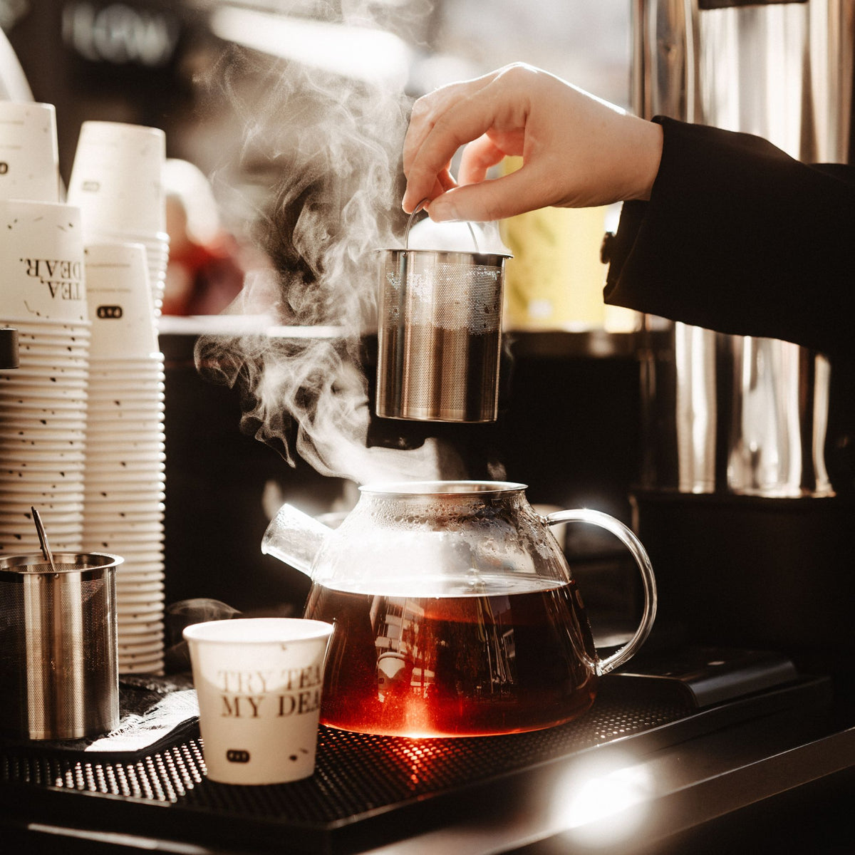 Glaskanne mit dampfendem, rotbraunem Tee und Metallsieb, weiße Becher mit TEA, DEAR. Branding