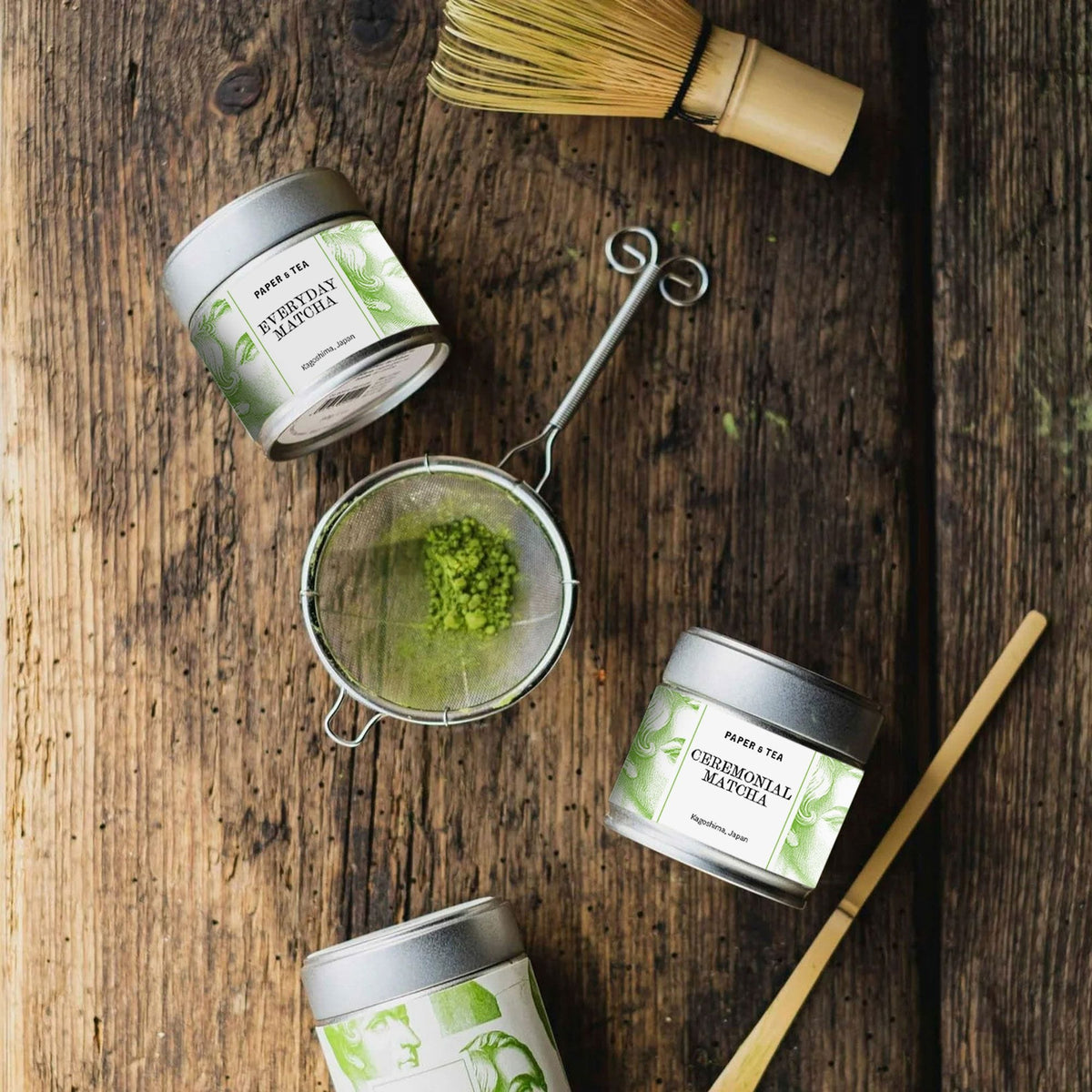 Drei Matcha-Metalldosen von PAPER & TEA aus Kagoshima mit Metallsieb und grünem Matcha-Pulver auf Holztisch.