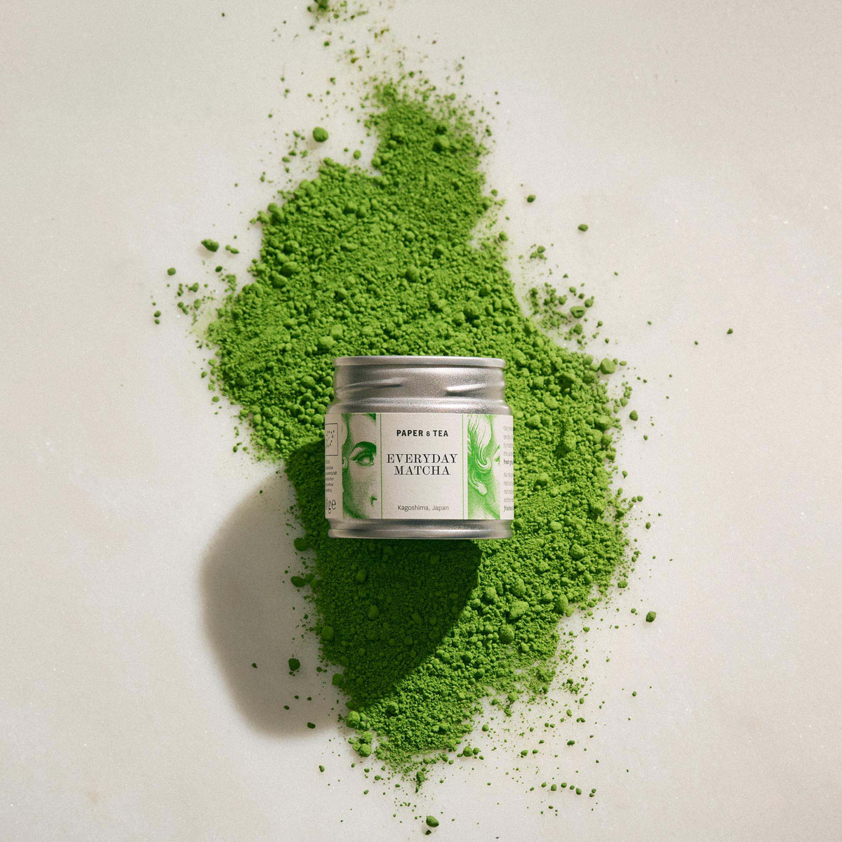 Silberne Dose Paper & Tea Matcha aus Kagoshima, Japan, umgeben von grünem Matcha-Pulver auf hellem Untergrund.