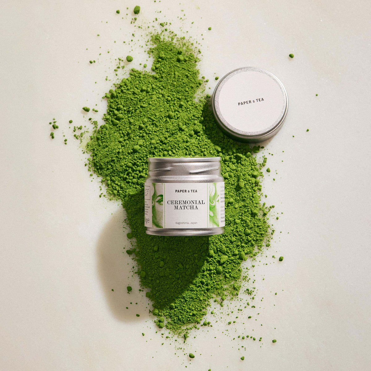 Kleine metallene Paper & Tea Matcha-Dose auf verstreutem, leuchtend grünem Matcha-Pulver aus Kagoshima, Japan