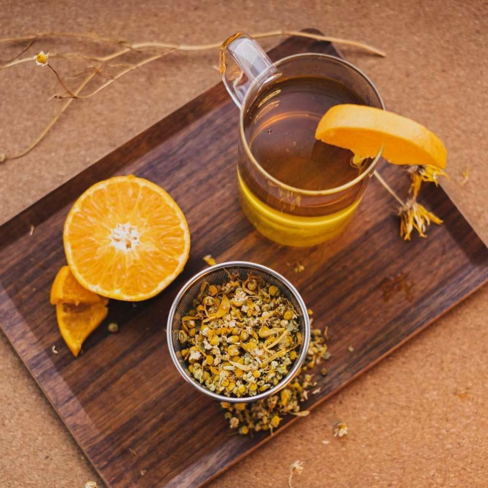 Transparentes Glas mit Kamillentee und Orangenscheibe auf dunklem Holzbrett neben halber Orange und Schale mit getrockneten Kamillenblüten.