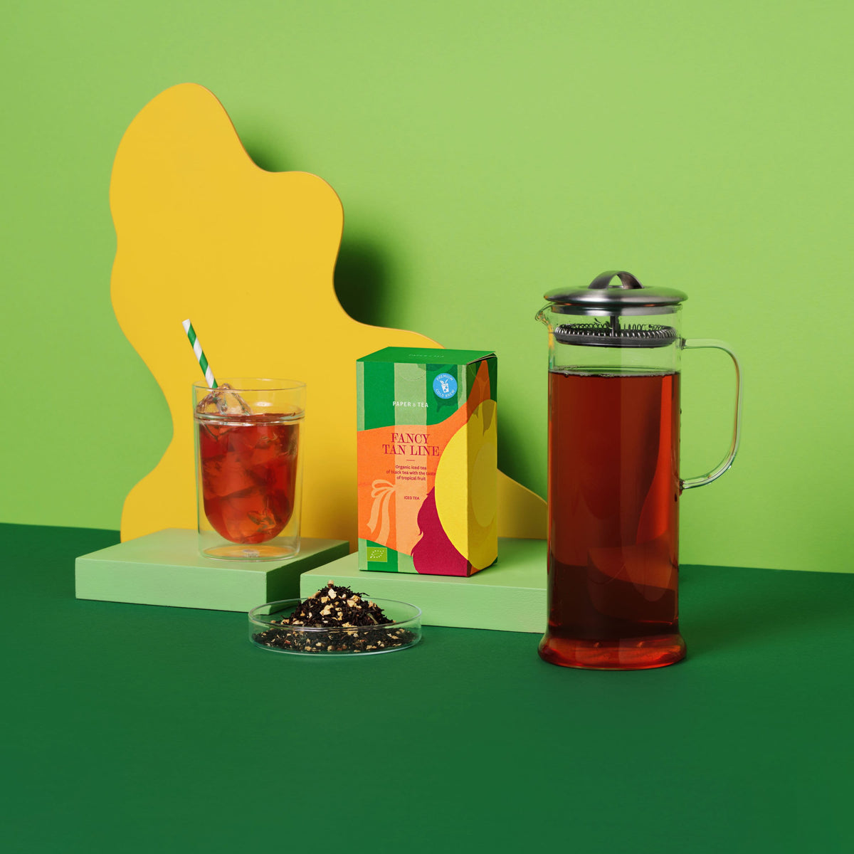 PAPER & TEA FANCY TAN LINE Bio-Eistee neben Glas mit Eis, French Press und losem Schwarztee vor grünem Hintergrund.