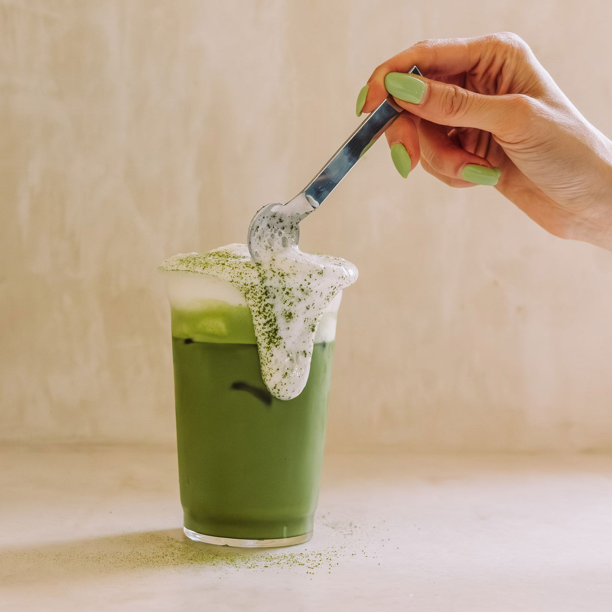 Klares Glas mit grünem Eistee und schaumiger Matcha-Schicht, Hand mit grünen Nägeln schöpft Schaum mit Löffel.