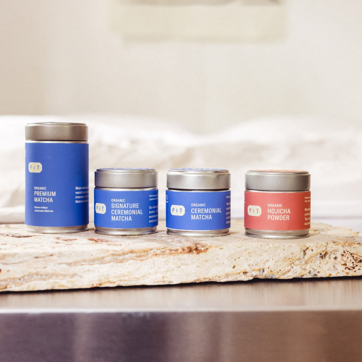 Vier P&T Teedosen auf Steinfläche: große blaue Dose Premium Matcha, zwei kleinere blaue Ceremonial Matcha und eine rote Hojicha Powder Dose.