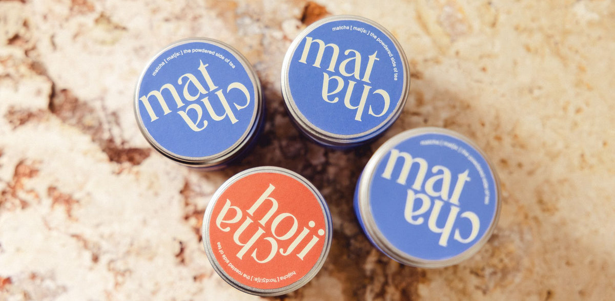Vier zylindrische Teedosen mit blauen Matcha-Deckeln und einem orangefarbenen Hojicha-Deckel auf beigem Steinhintergrund.