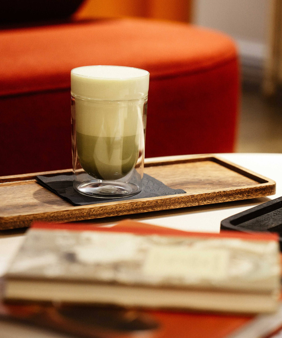 Glas mit zweischichtiger Matcha-Latte auf schwarzer Serviette im Holztablett vor rotem Sofa.