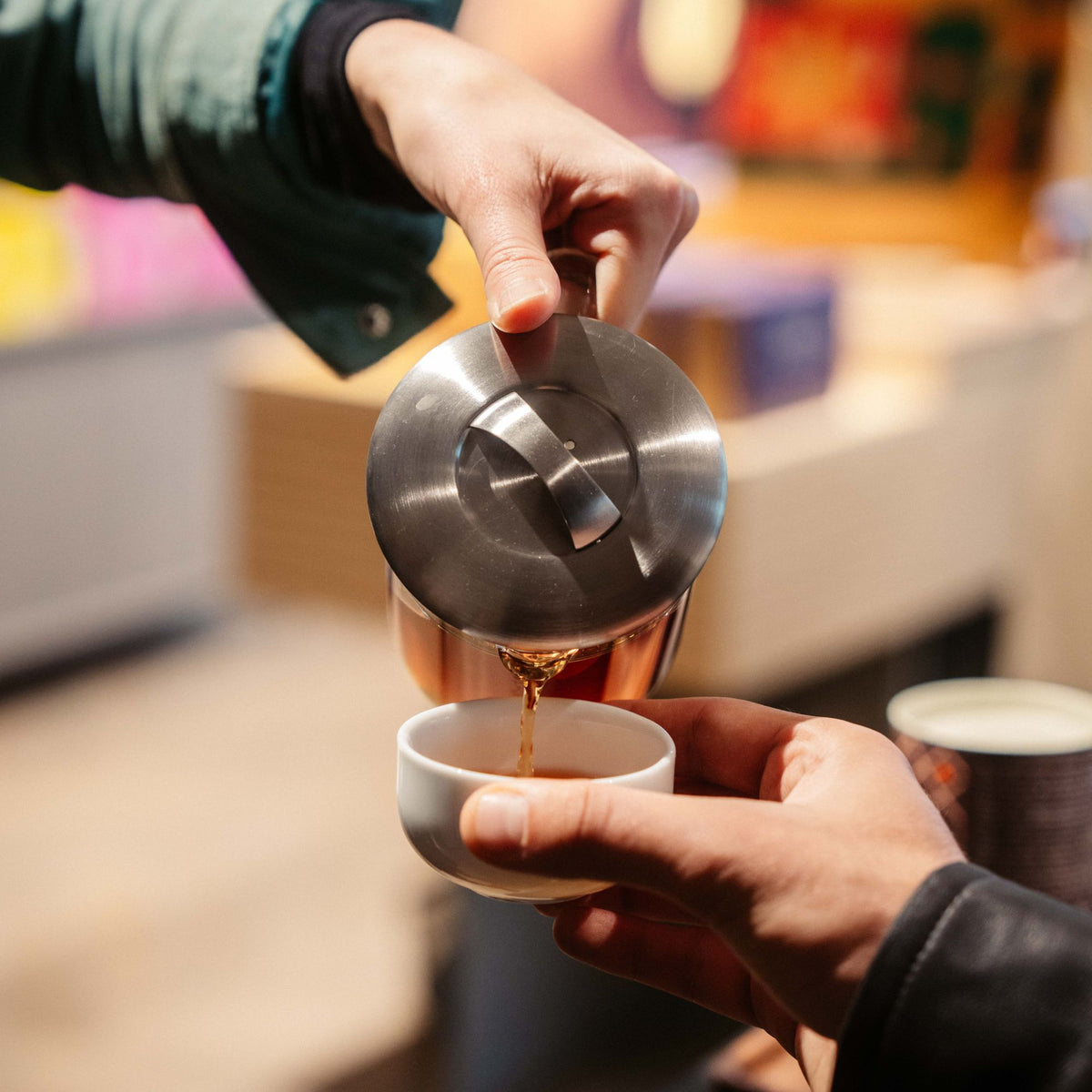 Hand gießt Tee aus einer Edelstahl-Teekanne in eine kleine weiße Keramiktasse.