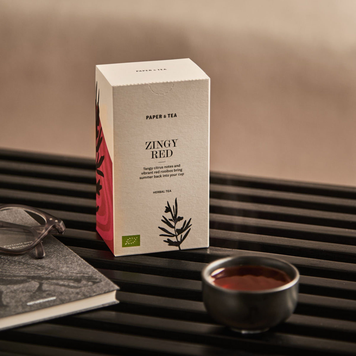 Paper & Tea Zingy Red Kräutertee-Verpackung neben roter Rooibos-Tasse, Buch und Brille auf dunklem Holz.