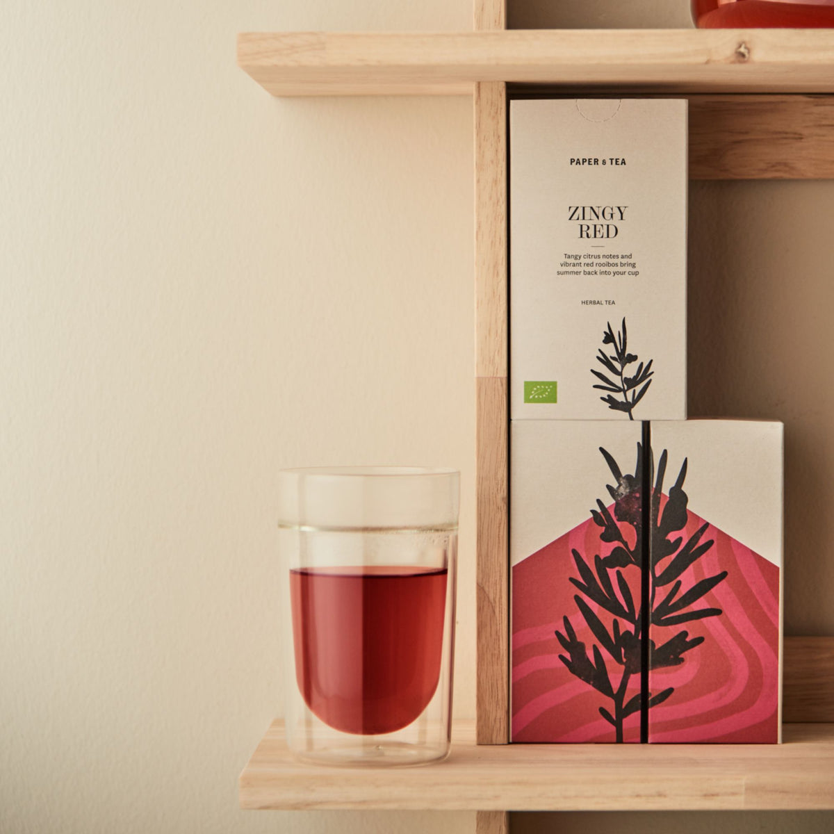 Zwei minimalistische Teeboxen für Zingy Red und ein Glas mit Kräutertee auf Holzregal.