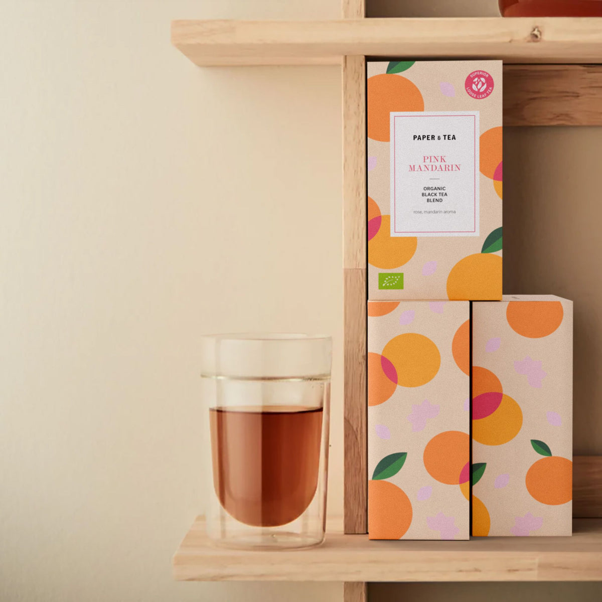 Holzregal mit PAPER & TEA Pink Mandarin Bio-Schwarztee-Boxen und Doppelwand-Glas mit Tee.