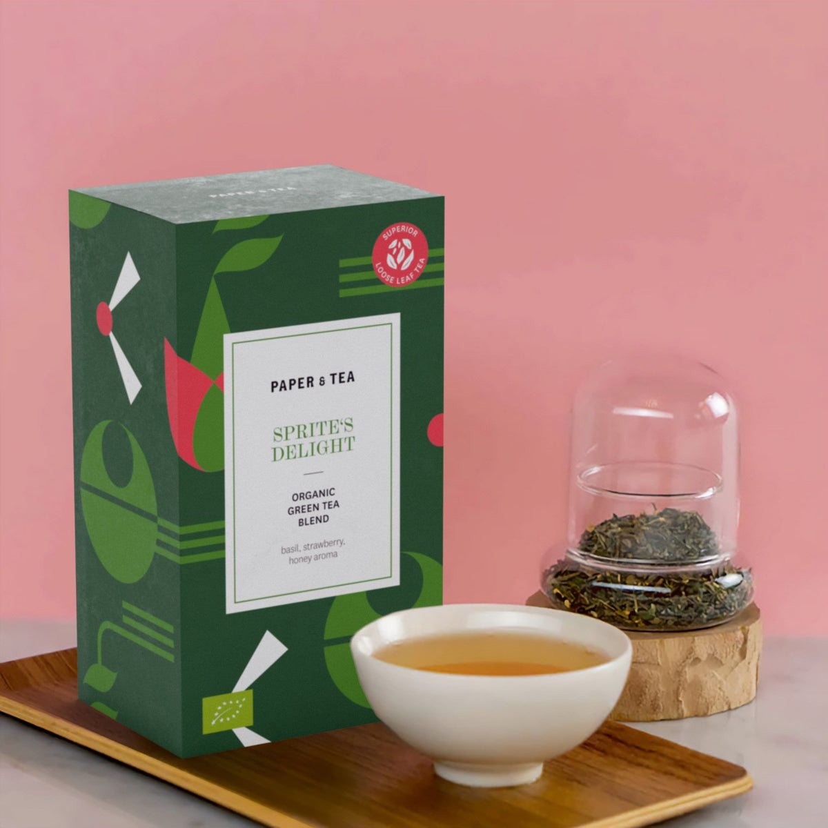 Grüne Geschenkbox mit Bio-Grüntee 'Sprite's Delight', daneben aufgebrühter Tee und Glasglocke auf Holztablett.