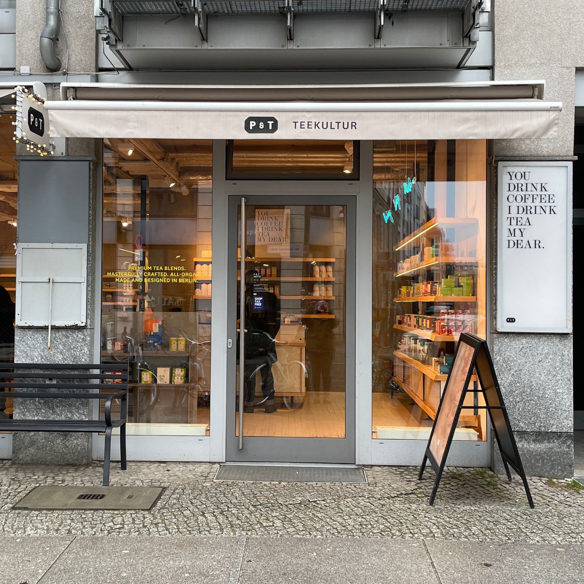 P & T TEEKULTUR Teehaus mit beleuchteten Regalen und 'YOU DRINK COFFEE I DRINK TEA MY DEAR.' Schriftzug.