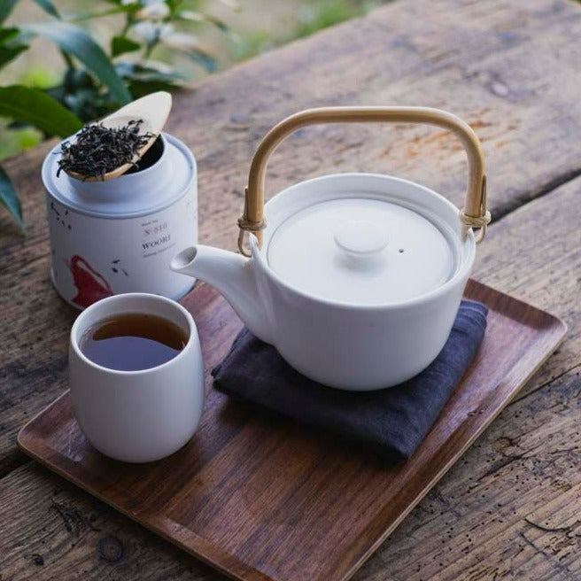 Weiße Keramik-Teekanne mit Bambusgriff und Tasse auf Holztablett neben Dose WOOR-Tee auf Holztisch