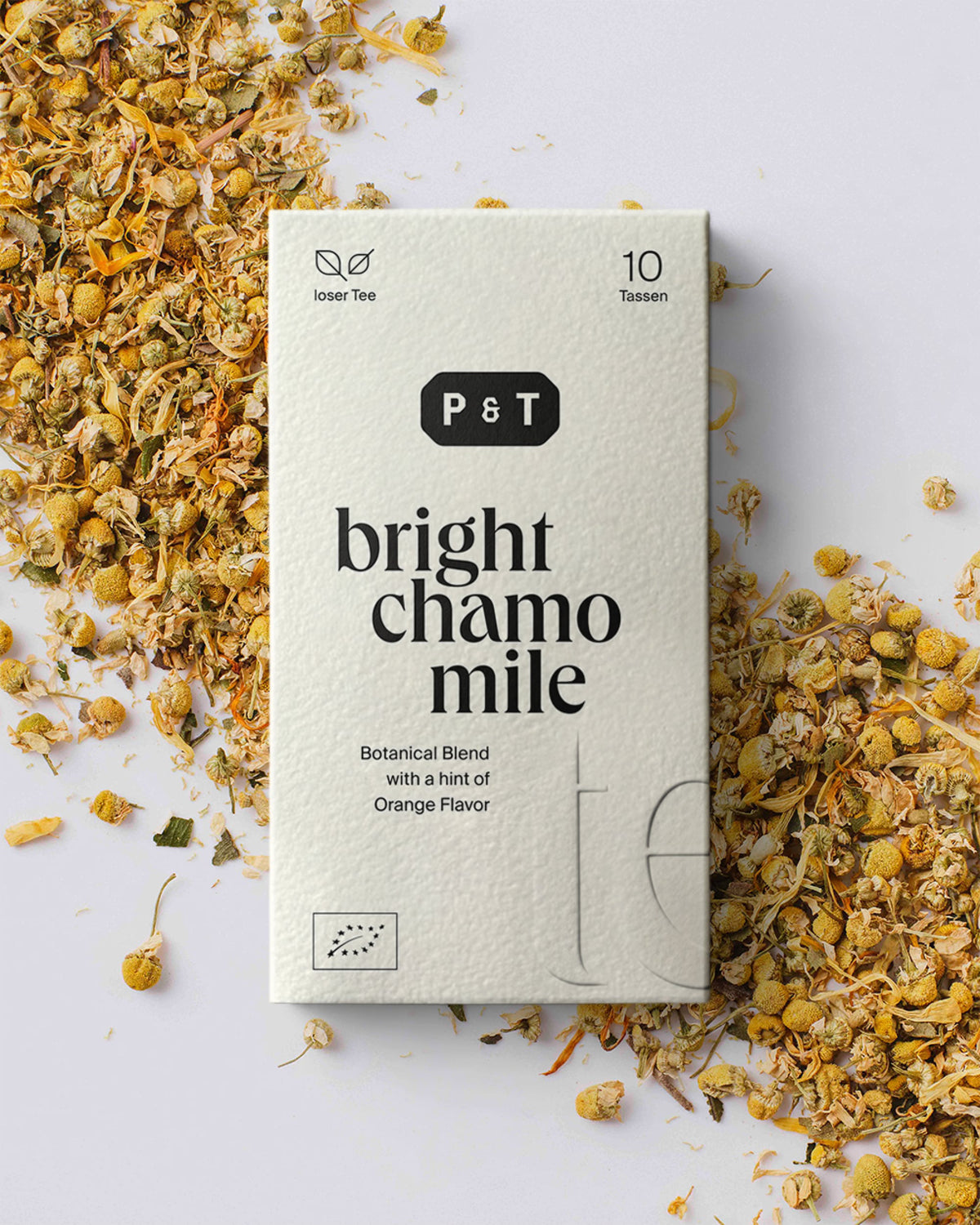 Weißer P&T-Teekarton mit Bright Chamomile-Kräutermischung und losen Kamillenblüten auf hellem Untergrund