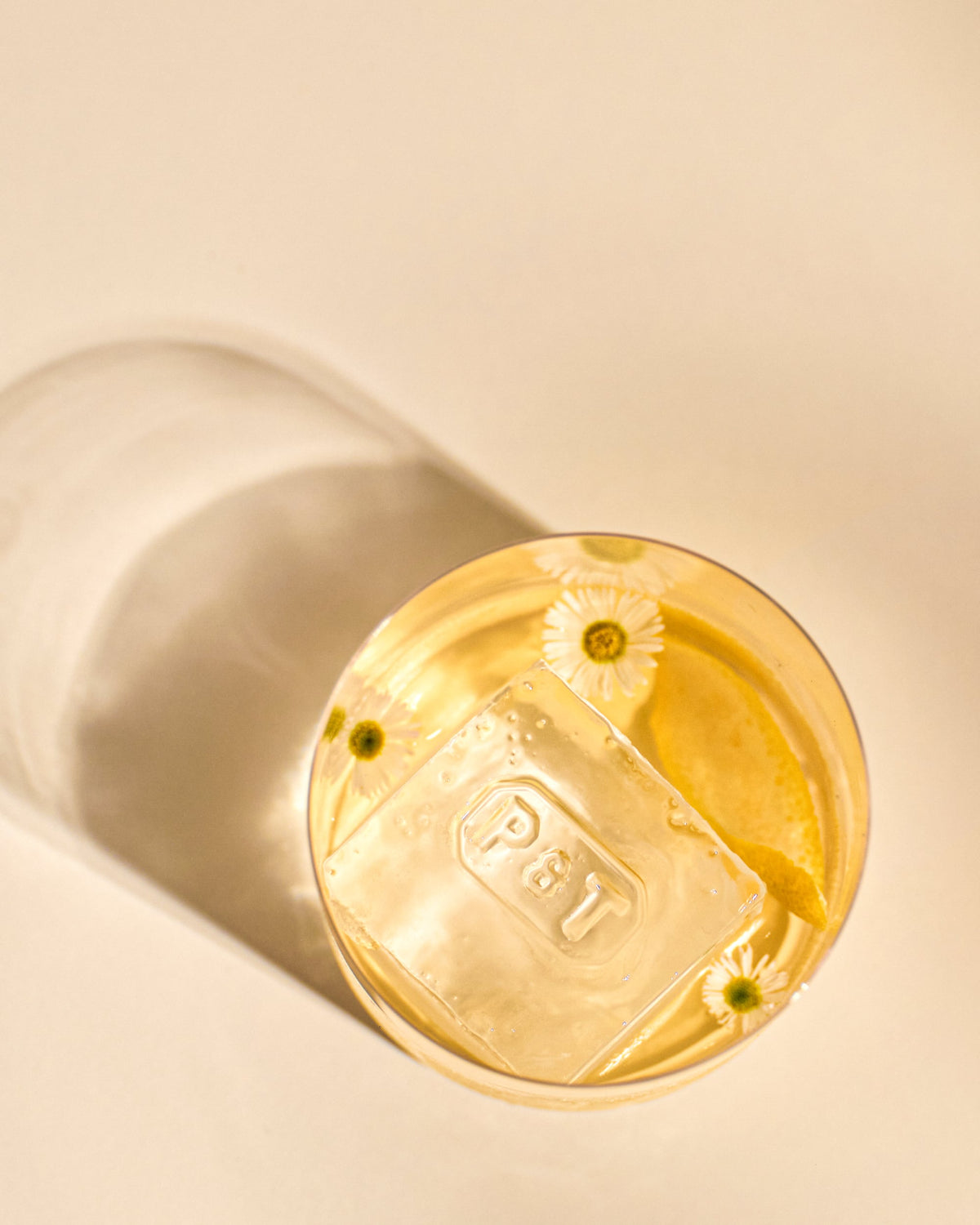 Rundes Glas mit zartgelbem Tee, großem Eiswürfel mit P&T-Prägung, Zitronenschale und Gänseblümchen.