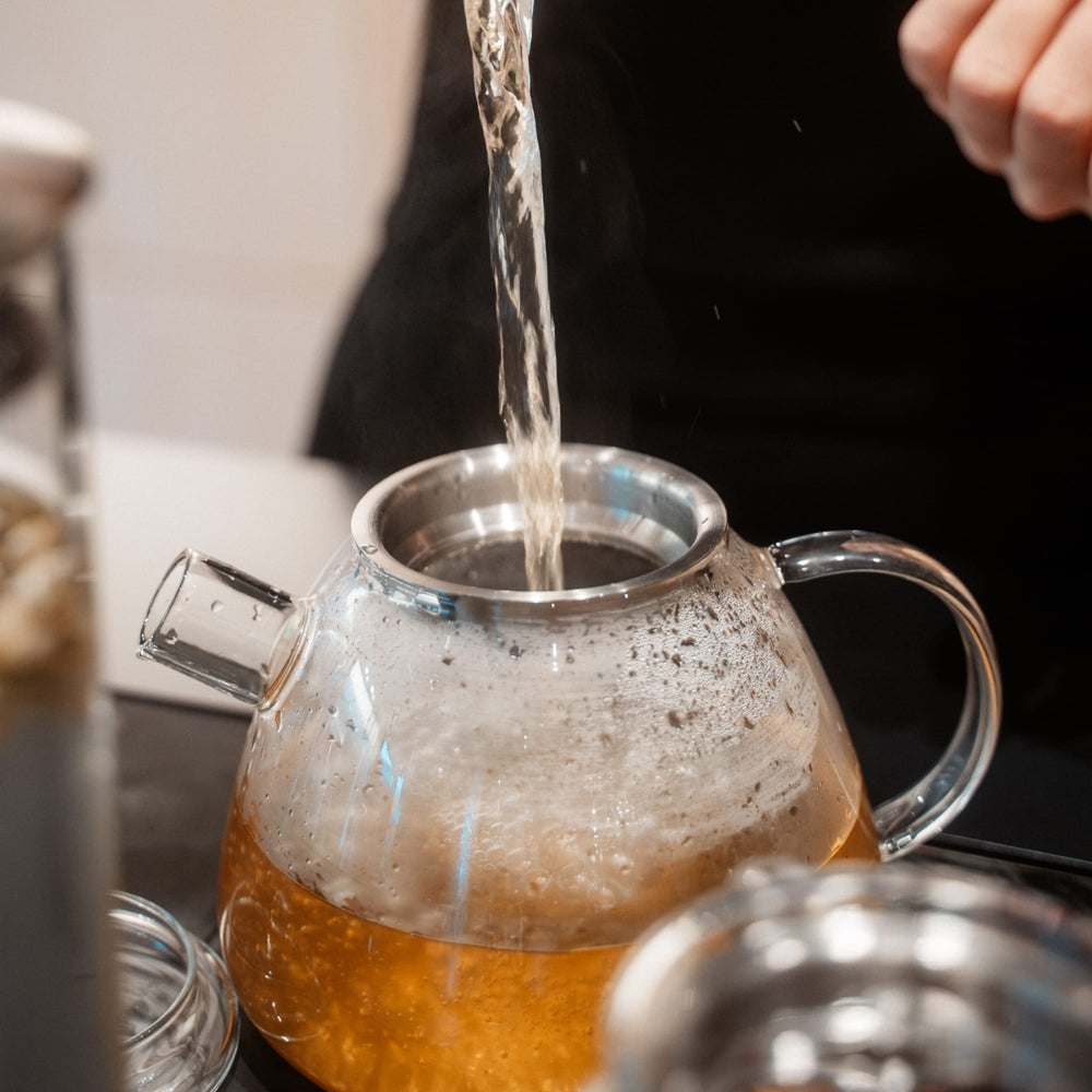Transparenter Glas-Teekessel mit Metallrand wird mit heißem Wasser gefüllt, Tee zieht.