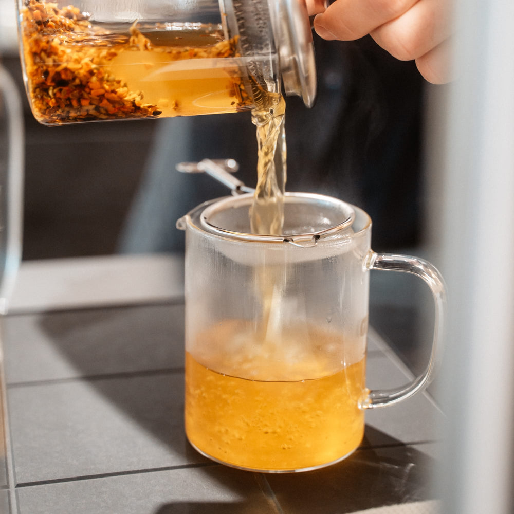 Glaskrug mit losen Teeblättern gießt orangefarbenen Tee in Glastasse durch Edelstahl-Teesieb auf Kacheluntergrund.