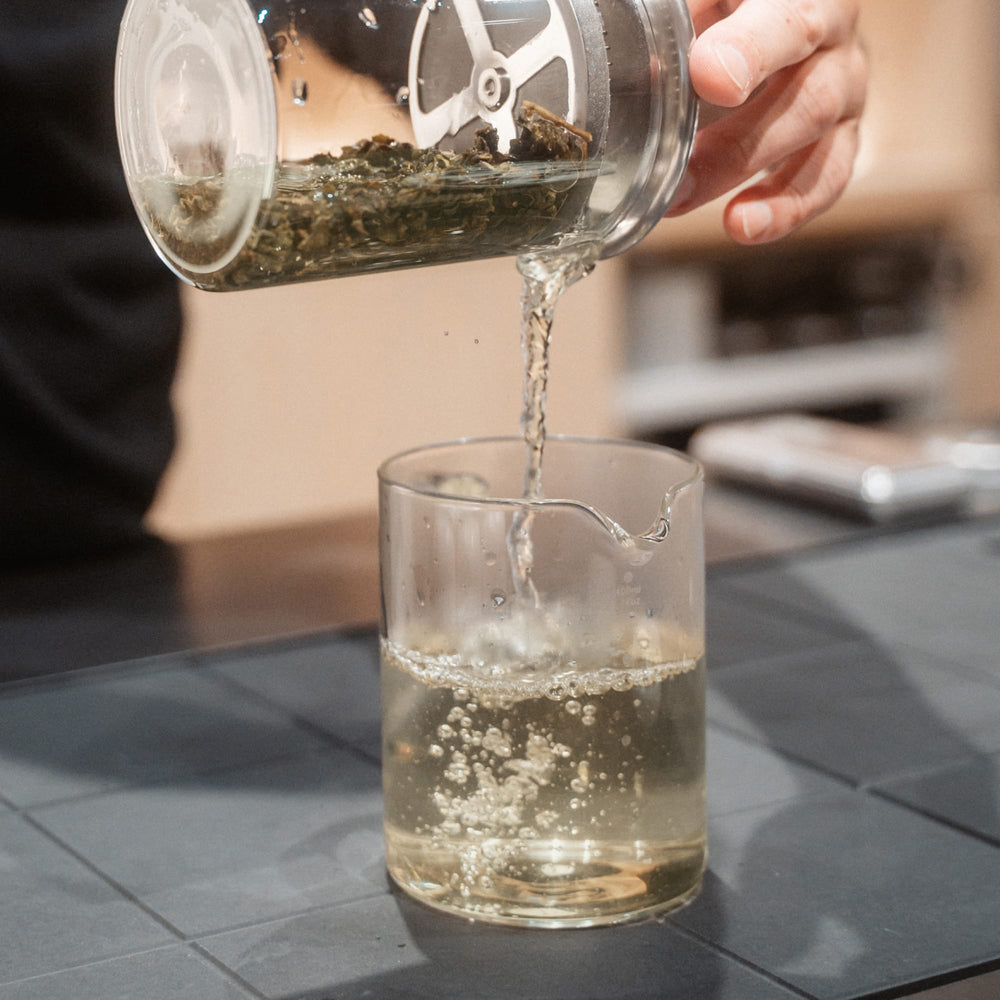 Frisch aufgebrühter loser Tee wird aus einem Glasbehälter in ein klares Becherglas gegossen.
