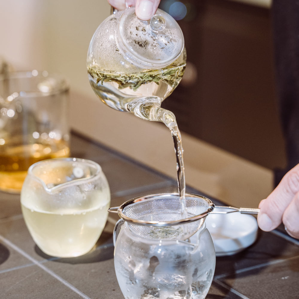 Grüner Tee fließt aus Glaskrug durch Teesieb in Glas-Tasse, Teezeremonie auf gefliestem Tisch.