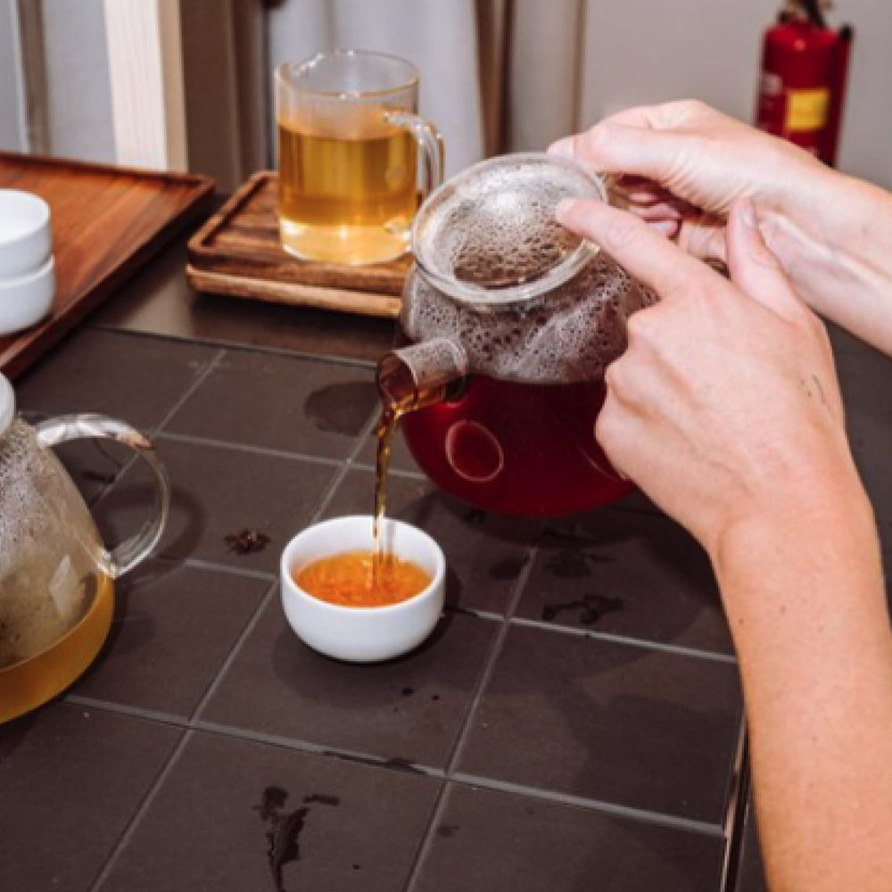 Person gießt Tee aus Glas-Teekännchen in weiße Tasse auf dunklem Fliesentisch