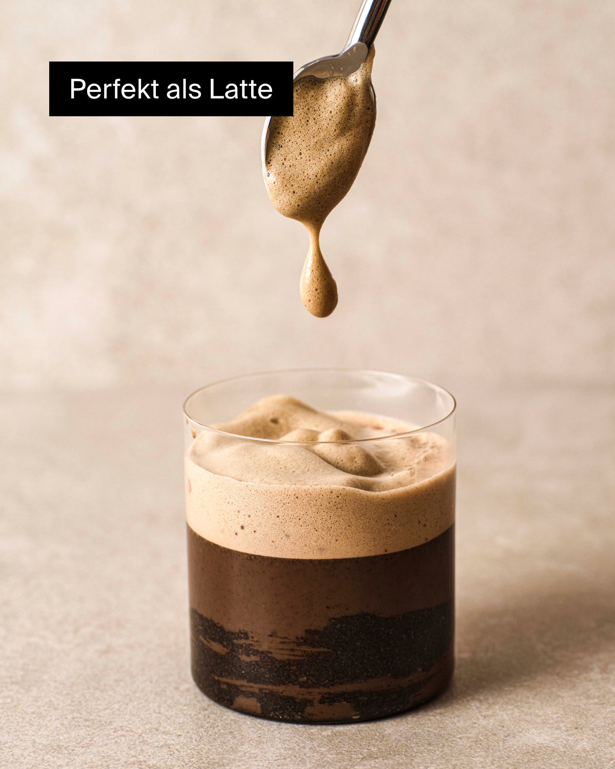 Transparenter Glasbecher mit Schichtkaffee, unten dunkel und oben cremiger Schaum, daneben ein Löffel.