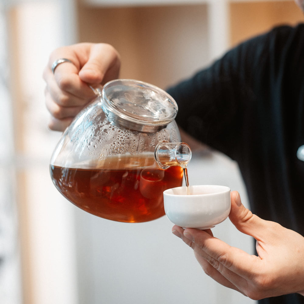 Glasteekanne mit dunkelbernsteinfarbenem Tee, gießt Tee in weiße Keramiktasse.