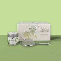 Grünes und weißes SATTVA Lemon Tea Blend Set mit botanischen Illustrationen, Teeglas und Metallsieb vor grünem Hintergrund.