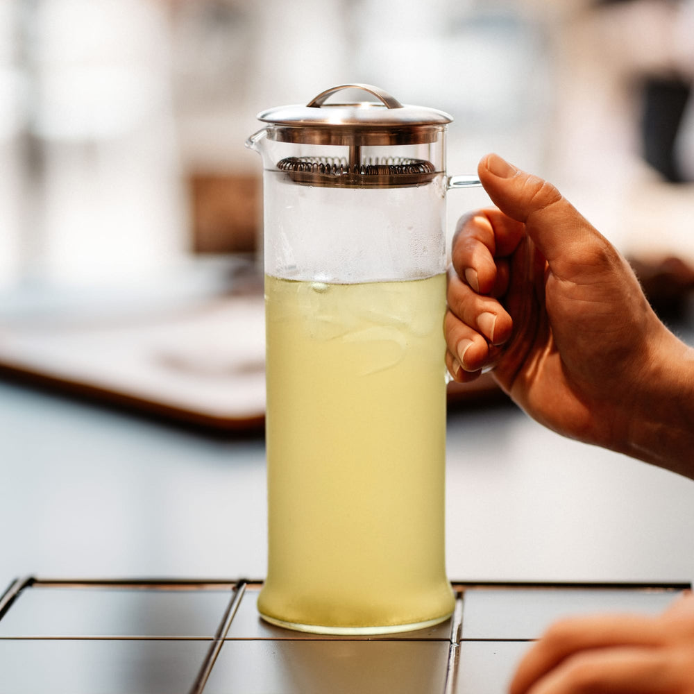 Hand hält transparente French Press mit eisgekühltem grünen Tee vor unscharfem Hintergrund.