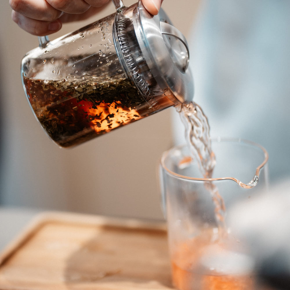 Hand gießt frisch aufgebrühten Tee aus einem transparenten French Press in ein Glas auf einem Holztablett.