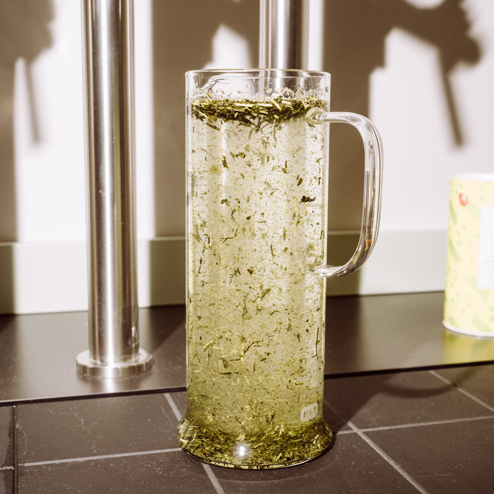 Transparente Glastasse mit hellem Grüntee und schwimmenden Blättern vor Edelstahlrohren und Behälter