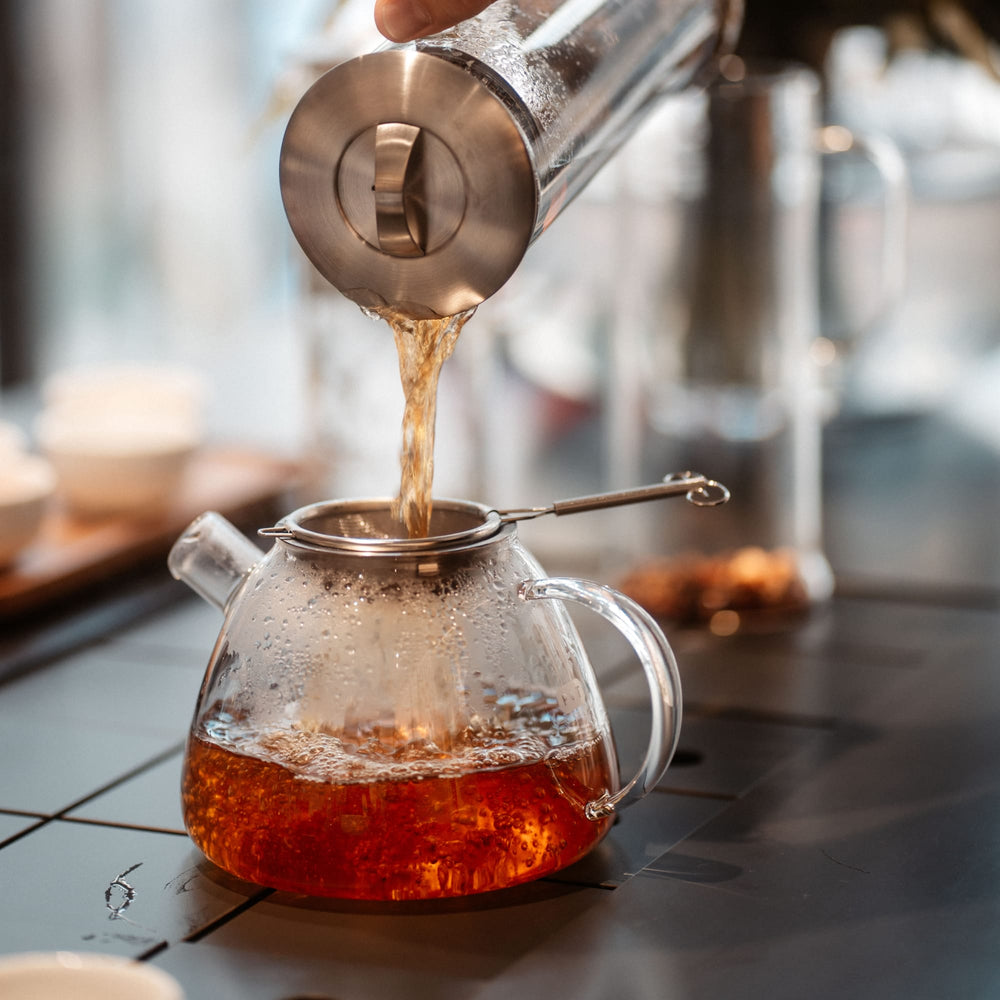 Glasteekanne mit bernsteinfarbenem Tee, der durch ein Metallsieb in eine Tasse gegossen wird.