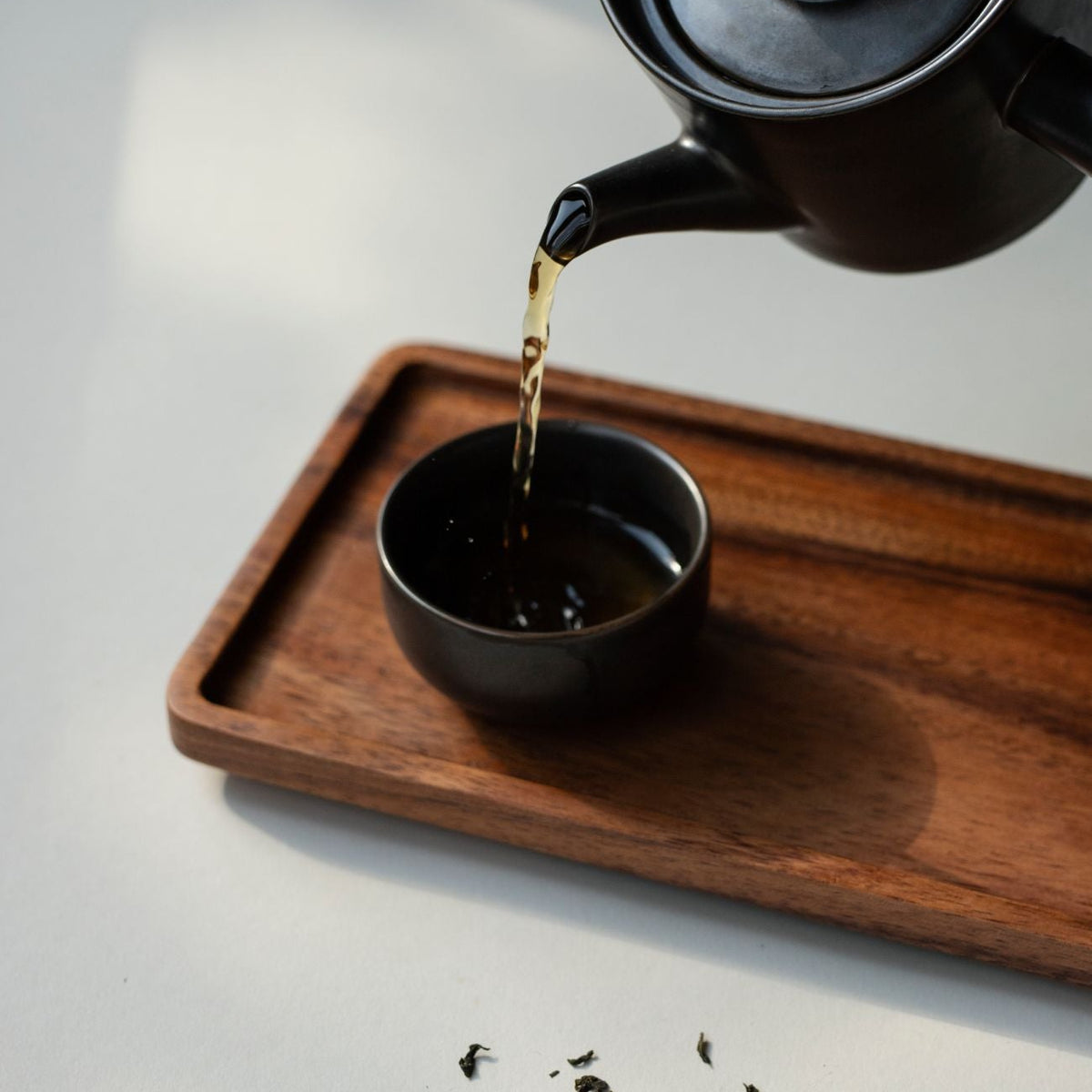 Schwarze Keramik-Teekanne gießt Tee in kleine Tasse auf Holztablett, umgeben von losen Teeblättern.
