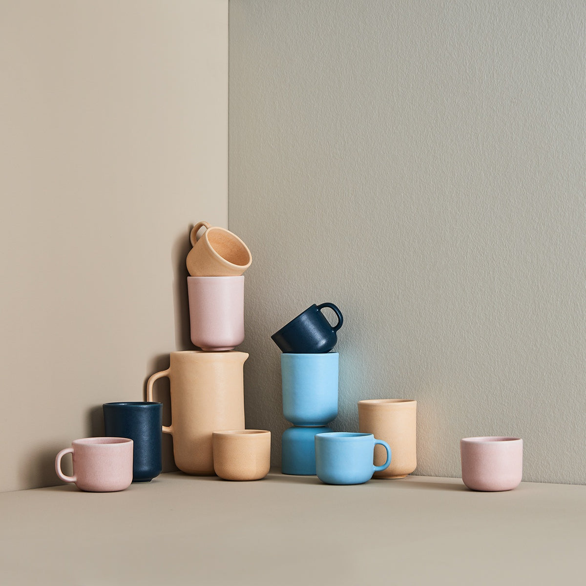 Zwölf matte Keramiktassen in Pastellfarben wie Rosa, Blau und Beige, kunstvoll an beiger Wand angeordnet.