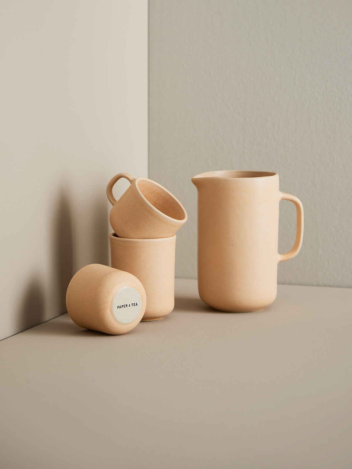 Beiges minimalistisches Keramik-Teeset mit einer Kanne und drei kleinen Tassen, eine Tasse gekippt mit Schriftzug PAPER & TEA.