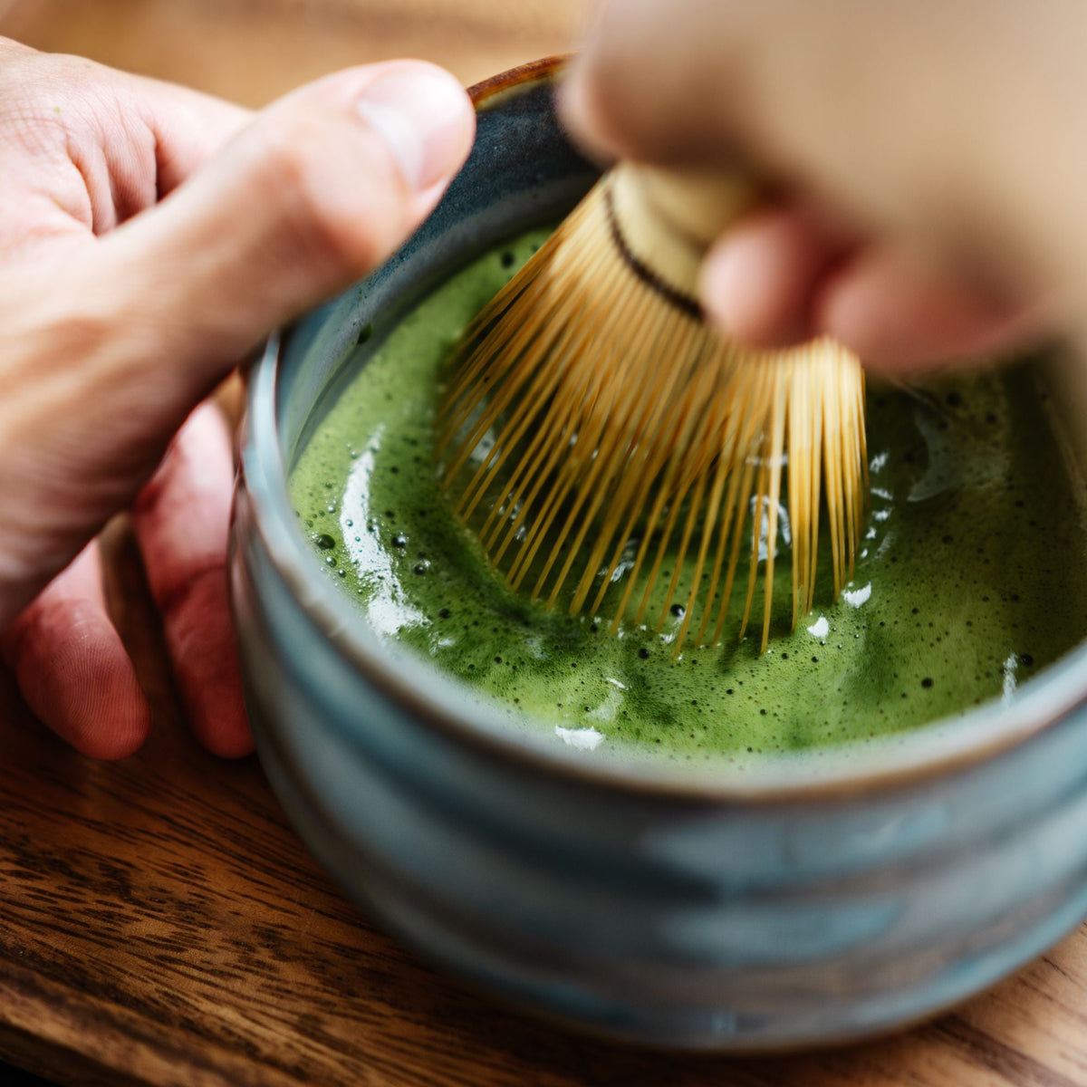 Hände bereiten cremigen grünen Matcha-Tee in einer Keramikschale mit traditionellem Bambusbesen zu.