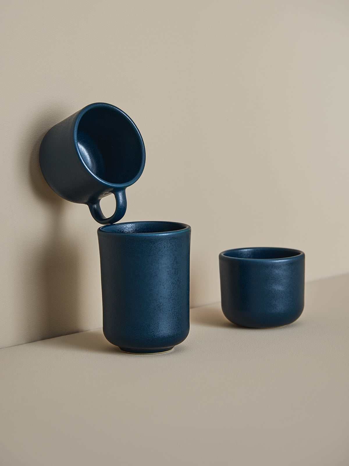 Drei dunkelblaue Keramiktassen vor beigem Hintergrund: hängend, hoch und rund, minimalistisch und elegant.