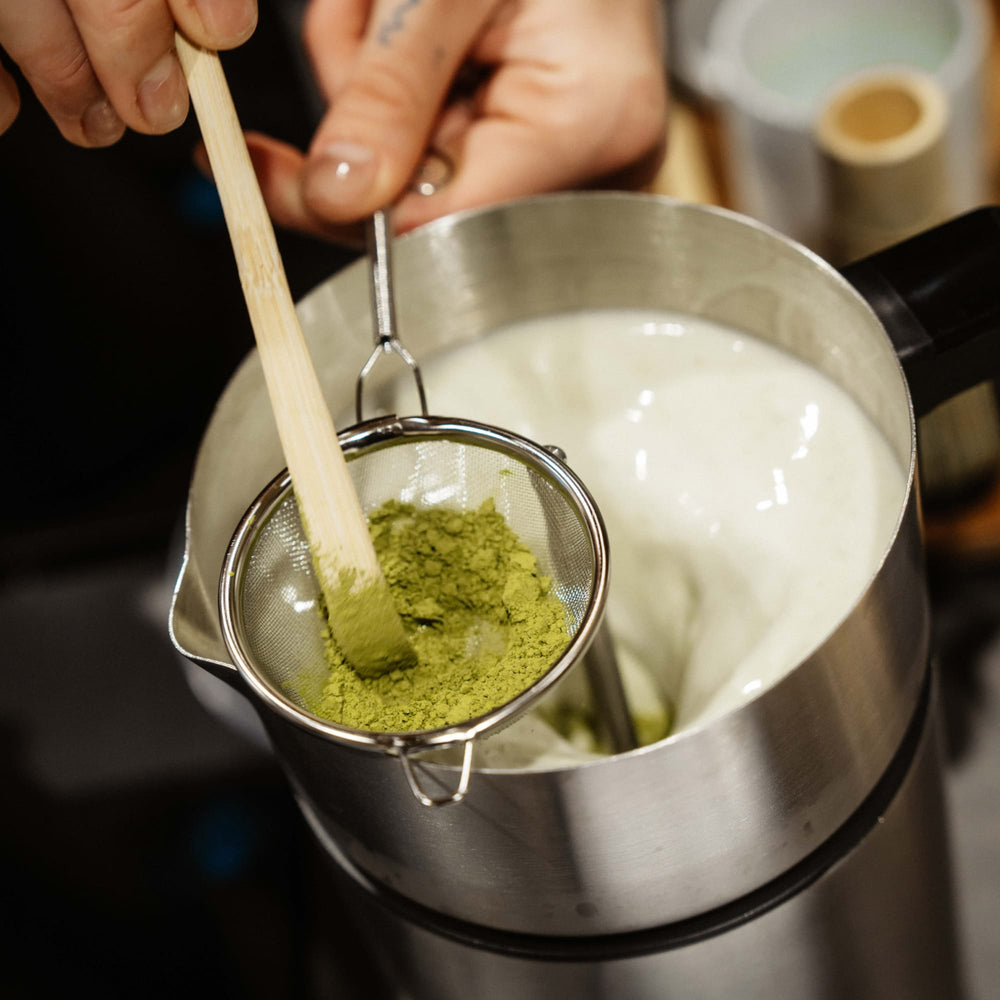 Hand hält Bambusschaufel mit grünem Matcha-Pulver über Edelstahlkanne mit schaumiger Milch und Teesieb.