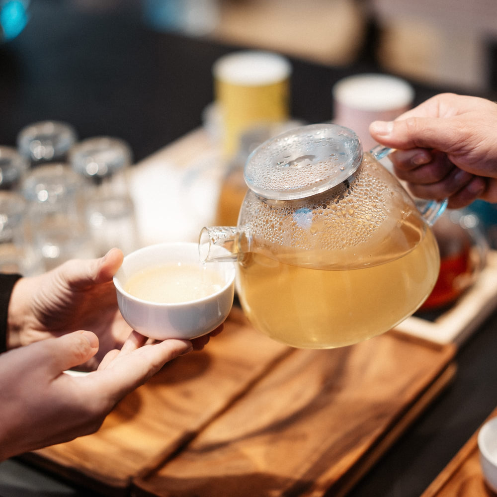 Glasteekanne mit hellem Tee gießt in weiße Keramiktasse auf Holztisch, bunte Gläser im Hintergrund