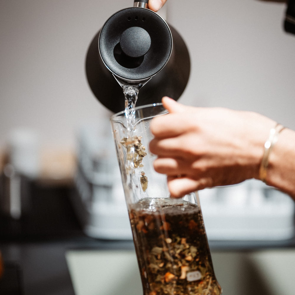 Ein schwarzer Wasserkocher gießt heißes Wasser in ein Glas mit Teeblättern, gehalten von einer Hand mit Armband.
