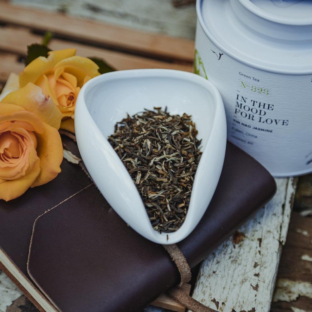 Weißes, tropfenförmiges Keramikgefäß mit YIN HAO JASMINE Grüntee auf braunem Notizbuch neben zwei gelb-orangen Rosen.