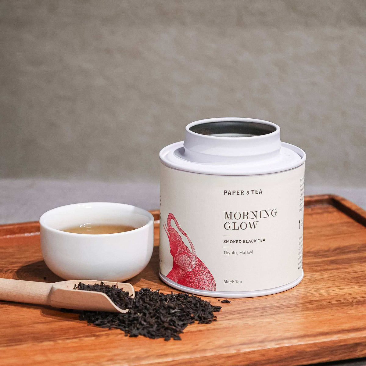 Weißer Papierbehälter mit Tee 'Morning Glow Smoked Black', rote Elefanten, Keramiktasse, Holztablett.