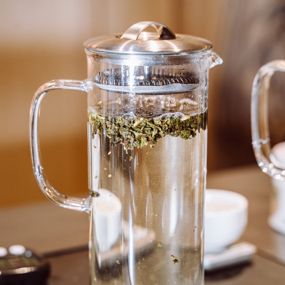 Glaskaraffe mit grünem Tee, im Hintergrund unscharfe weiße Keramiktasse.