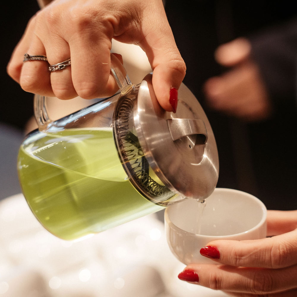 Hand mit roten Nägeln gießt hellgrünen Tee aus einer Glasteekanne in eine weiße Tasse.