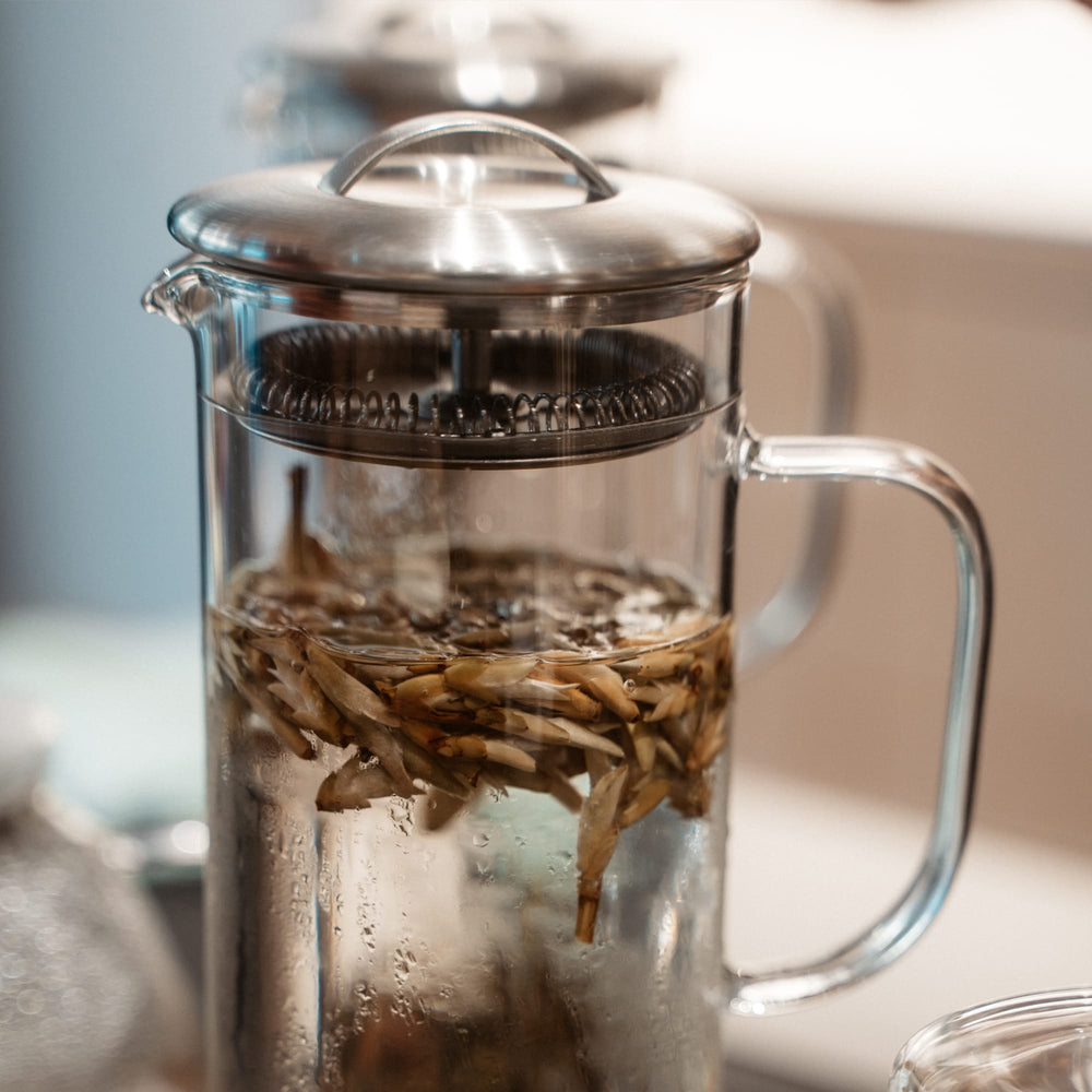 Transparente Glas-French-Press mit Wasser und Teeblättern, glänzender Metalldeckel und Stempel.