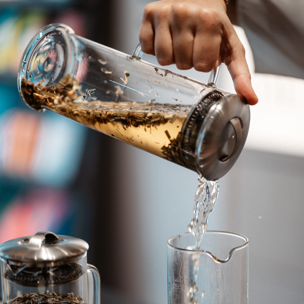 Hand gießt frisch gebrühten Tee mit Teeblättern aus Glaskanne in Glas bei beruhigendem Licht.