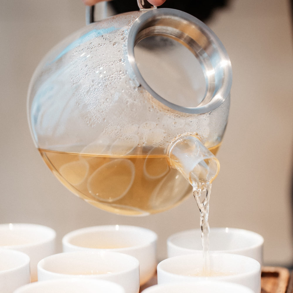 Transparente Glaskanne mit Metallrand gießt hellbraunen Tee in weiße Keramiktassen.