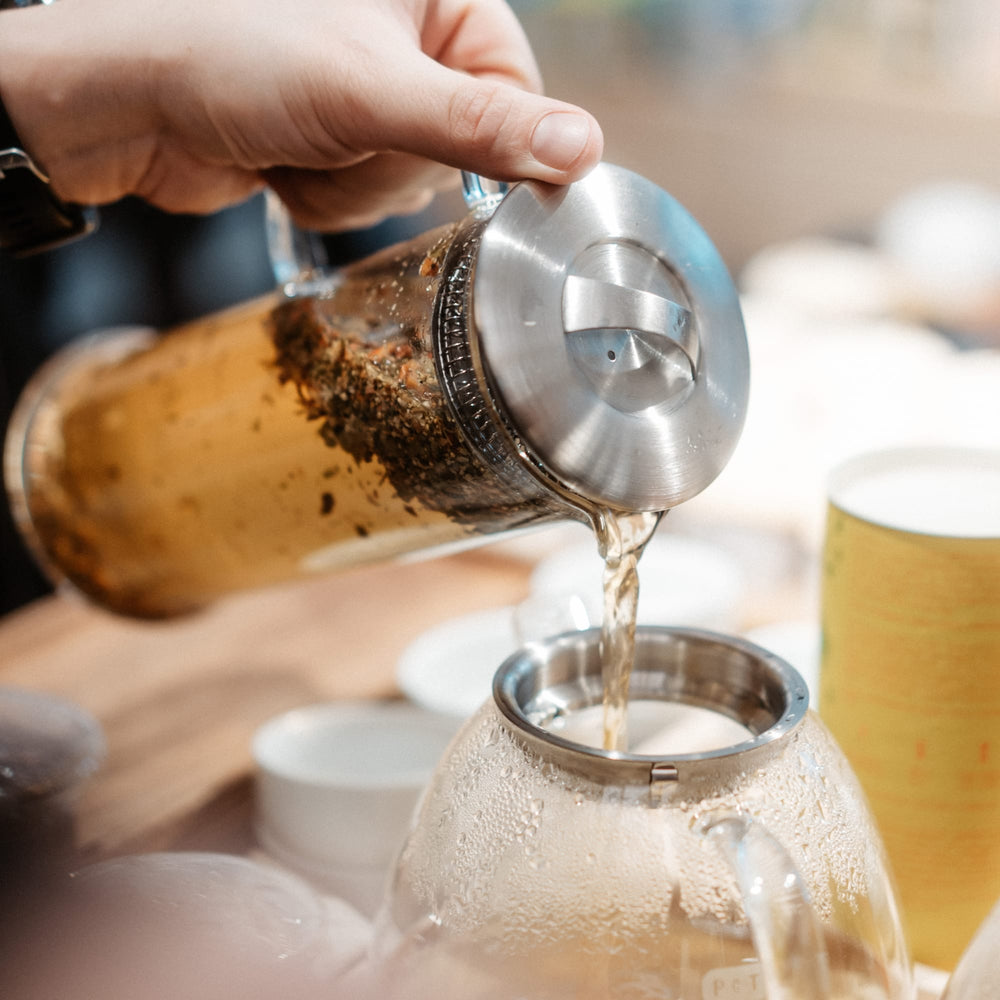 Hand gießt heißen Tee aus French Press in gläserne Teekanne, Tassen und gelbe Teedose im Hintergrund.