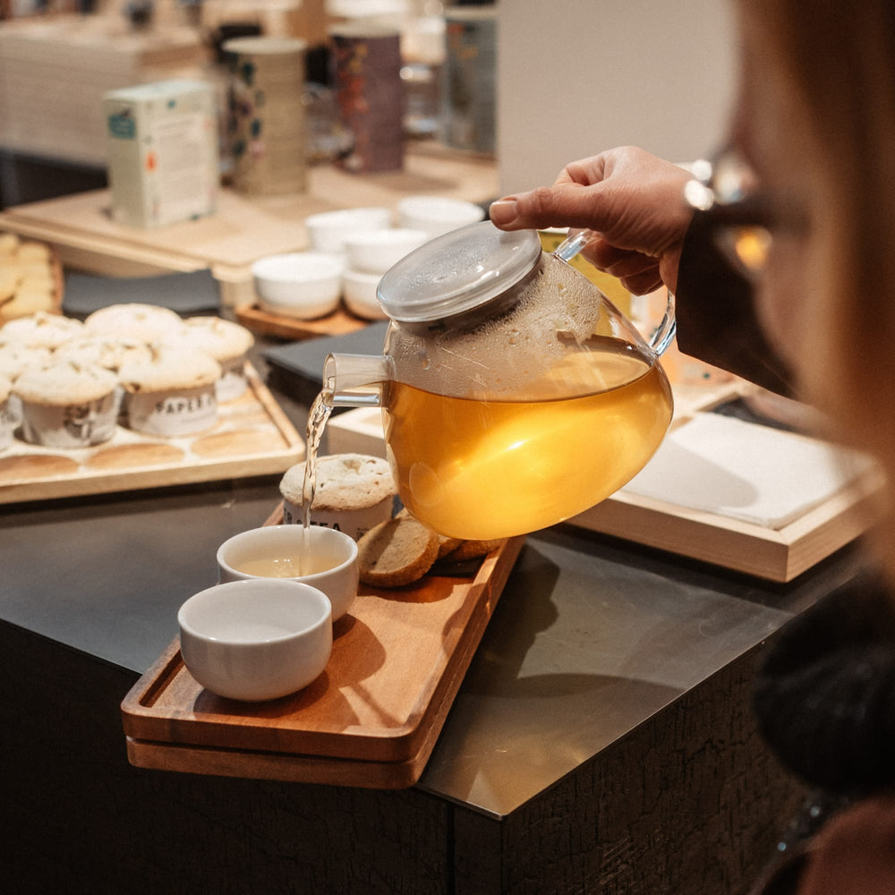 Hand gießt bernsteinfarbenen Tee aus Glaskanne in weiße Tasse auf Holztablett mit Keksen.